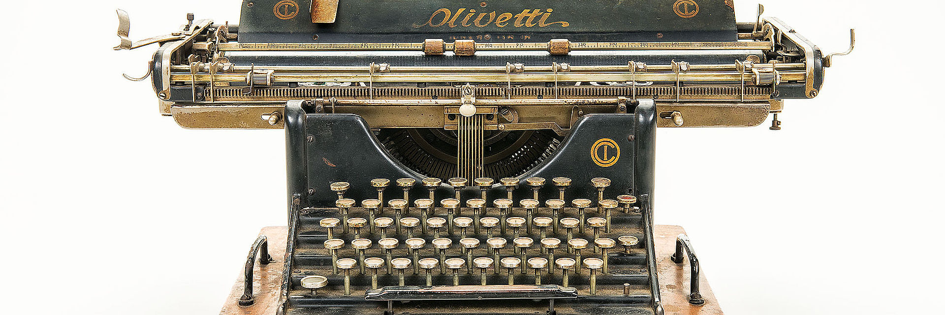 Farbfoto mit Frontalansicht der großen Schreibmaschine Olivetti M 20. Sie ist matt schwarz, besitzt wenig schmückende Elemente und ist auf ein massives Holzbrett geschraubt. Sie weist starke Gebrauchsspuren auf. 