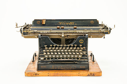 Farbfoto mit Frontalansicht der großen Schreibmaschine Olivetti M 20. Sie ist matt schwarz, besitzt wenig schmückende Elemente und ist auf ein massives Holzbrett geschraubt. Sie weist starke Gebrauchsspuren auf. 