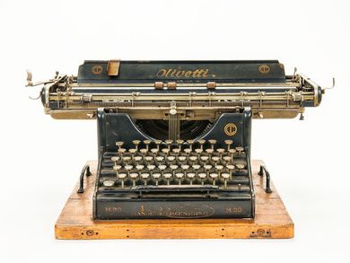 Farbfoto mit Frontalansicht der großen Schreibmaschine Olivetti M 20. Sie ist matt schwarz, besitzt wenig schmückende Elemente und ist auf ein massives Holzbrett geschraubt. Sie weist starke Gebrauchsspuren auf. 