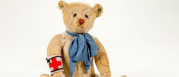 Zu sehen ist ein beiger Teddybär mit offensichtlich kaputten und abgewetzten Stellen. Er trägt eine blaue Schleife um den Hals und eine weiße Armbinde mit rotem Kreuz.