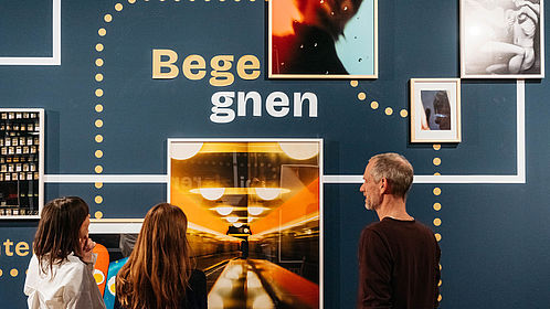 An einer dunkelblauen Wand hängen mehrere gerahmte Bilder. In der Mitte ist das Wort "Begegnen" zu lesen. Zwischen den einzelnen Fotografien sind gepunktete Verbindungen und Verbindungslinien zu sehen. Drei Personen stehen vor den Bildern und betrachten diese.