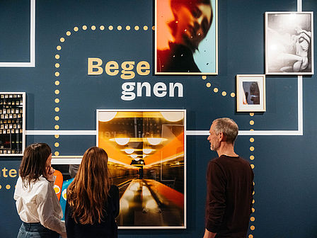 An einer dunkelblauen Wand hängen mehrere gerahmte Bilder. In der Mitte ist das Wort "Begegnen" zu lesen. Zwischen den einzelnen Fotografien sind gepunktete Verbindungen und Verbindungslinien zu sehen. Drei Personen stehen vor den Bildern und betrachten diese.