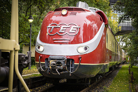 Ein roter Zug mit geschwungener Verzierung steht auf einem begrünten Gleisabschnitt.