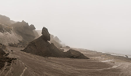 An einem Strand mit braunem Sand ragen dunkle Erdhügel in Richtung des Meeres. Sie sehen aus wie Eisberge auf dem Trockenen. Himmel und Meer sind gleichmäßig grau. Ein Horizont ist nicht zu erkennen.