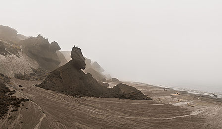 On a beach with brown sand, dark mounds of earth jut out toward the sea. They look like beached icebergs. The sky and sea are uniformly gray. There is no visible horizon.