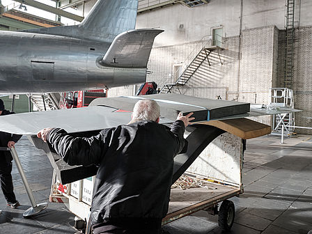 Ein älterer Herr hält eine Tragfläche eines Flugzeugs in den Händen, im Hintergrund ist der Rumpf des Flugzeugs zu sehen.
