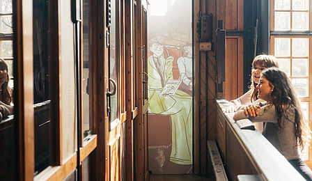 Zwei Frauen sehen auf einen Eisenbahnwagon, hinter ihnen ein Zeichentrickfilm.