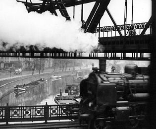Ein Schwarzweiß-Foto zeigt einen Kanal, über den eine Eisenbahnbrücke führt, außerdem sieht man S-Bahnschienen und eine Straße.