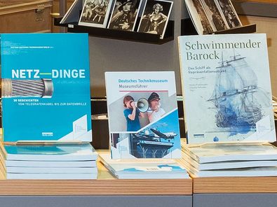 Auf einem Holztisch werden fünf verschiedene Publikationen präsentiert. Ein Buch steht jeweils aufrecht, weitere Exemplare liegen auf Stapeln davor.