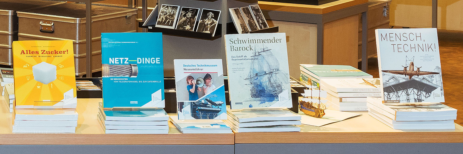 Auf einem Holztisch werden fünf verschiedene Publikationen präsentiert. Ein Buch steht jeweils aufrecht, weitere Exemplare liegen auf Stapeln davor.