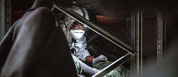 Eine Person mit dunkler Hautfarbe, in einen weißen Schutzanzug mit Mundschutz, Schutzbrille und Stirnlampe gekleidet, blickt durch eine Metallkonstruktion ins Bild. Im Vordergrund ist schemenhaft eine Figurine zu erkennen.