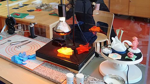 Auf einem Arbeitstisch werde mit einer Rotlichtlampe farbige Zuckerpaltten erwärmt. Rechts im Bild unter anderem ein Schwan und eine Rose aus Zucker geformt.