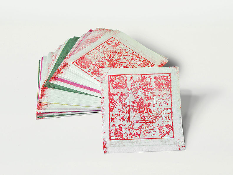 Ein Stapel quadratischer Glückspapiere, weiß mit rotem Aufdruck. Das oberste Papier ist heruntergenommen. Es zeigt Büsche, Bäume und Pflanzen.
