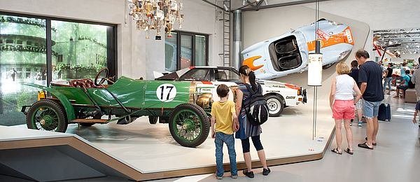 Blick in die Straßenverkehr-Ausstellung: Mehrere Personen stehen vor einem niedrigen Podest, auf dem zwei historische Rennwagen stehen. Ein weiterer blau-orangener Rennwagen ist an der Wand befestigt. Über dem Podest hängt eine Deckeninstallation aus zahlreichen Pokalen.