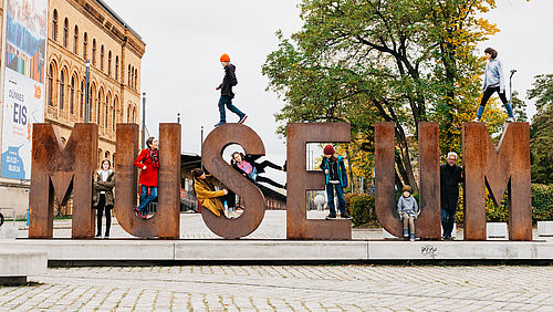 Auf der Wortskulptur "Museum" auf dem Vorplatz des Deutschen Technikmuseums haben sich Kinder und Erwachsene in und af die Buchstaben gesetzt.