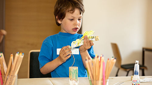 Ein kleiner Junge bastelt an einem Tisch. In seinen Händen hält er eine große Biene aus Papier. Vor ihm auf dem Tisch stehen eine Flasche Flüssigkleber und zwei Becher mit Buntstiften. 