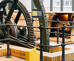 Vor einer Balancier-Dampfmaschine im Museum steht ein gelber Kanisterter und ein Objektschild.