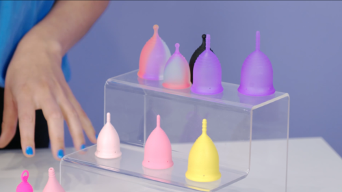 Abgebildet ist eine Hand neben verschieden großen, farbigen Menstruationstassen