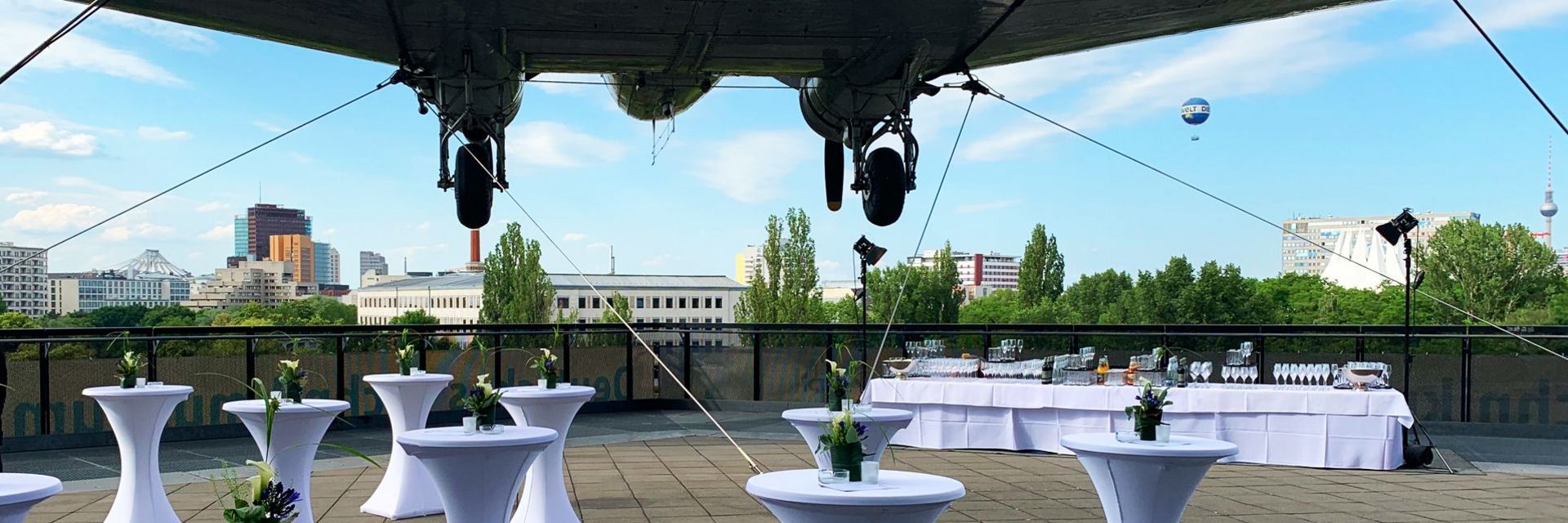Terrasse mit dekorierten Stehtischen über der ein Flugzeug hängt. Ausblick auf einen blauen Himmel, Gebäudedächer und den Berliner Fernsehturm.
