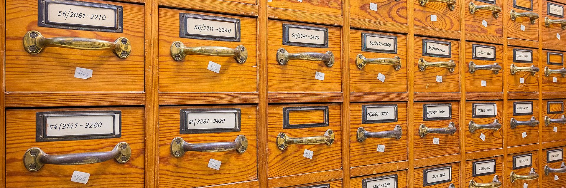 Ein historischer Archivschrank aus Holz mit vielen kleinen Schubladen mit Metallgriffen. Die Schubladen sind mit Signaturen versehen.