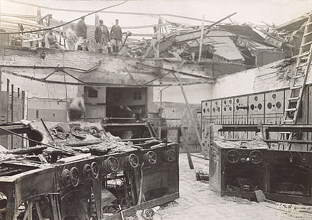 Das Foto zeigt Kriegsschäden am Kraftwerk Spandau 1943/1944. Im Vordergrund des Bildes sind zerstörte Bedienpulte mit Kurbelrädern zu sehen. Die Decke des Raumes ist zerstört. Im darüber liegenden Stockwerk stehen sechs Arbeiter. 