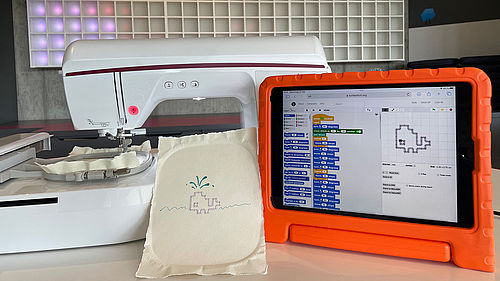 Ein Tablet mit Programmiercode steht neben einer Nähmaschine und einen Stück Stoff, auf das ein kleiner Wahl gestickt ist, den man auch auf dem Tablet sieht.