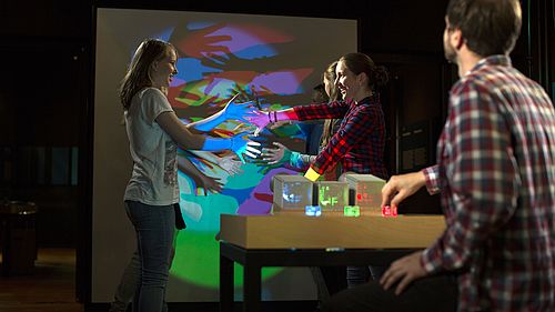 Drei junge Frauen beobachten, wie sie im Licht farbiger Scheinwerfer bunte Schatten werfen. Im Vordergrund bedient ein Mann die Scheinwerfer.