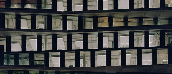Das Farbfoto zeigt den Teil einer Fassade eines modernen Bürogebäudes bei Nacht. Die Fenster sind unregelmäßig angeordnet, wodurch die Struktur wellenförmig erscheint. Helles Innenlicht beleuchtet die Arbeitsplätze und technischen Installationen. Die dunklen Außenflächen bilden dazu einen starken Kontrast.