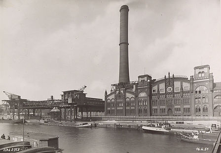 An der Spree befindet sich das Elektrizitätswerk Charlottenburg mit geschwungenen Formen und Rundbogenfenstern. In der Bildmitte ist ein großer Schornstein zu sehen. Die Krananlage für die Entladung der Kohleschiffe befindet sich links im Bild.