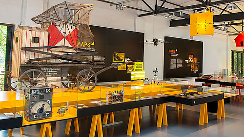 Der Ausstellungsbereich MAPS besteht aus einem sehr großen Tisch. Darauf befinden sich Objekte, Medienstationen, Texte und Grafiken. Über dem Tisch hängt das verkleinerte Modell einer Landkarte in Gelb.