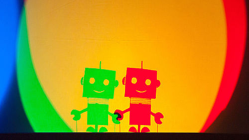 Auf einen dunklen Hintergrund fällt ein gelber Lichtkreis. Links am Rand des Kreises befindet sich ein grünes und rechts ein rotes Kreissegment. Am unteres Rand des Lichtkreises befinden sich zwei identische Roboter-Figuren nebeneinander, rechts in rot und links in grün. An der Hand überlappen sich beide Figuren. Der überlappende Teil ist schwarz. 