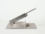 Der Stierhodenschneider wurde um 1950 von der Firma Schering gebaut. Es handelt sich um eine besonders stabile Schlagmesserklinge, die auf eine Metallplatte montiert ist, und mit einem Metallgriff an der Seite heruntergedrückt wird.