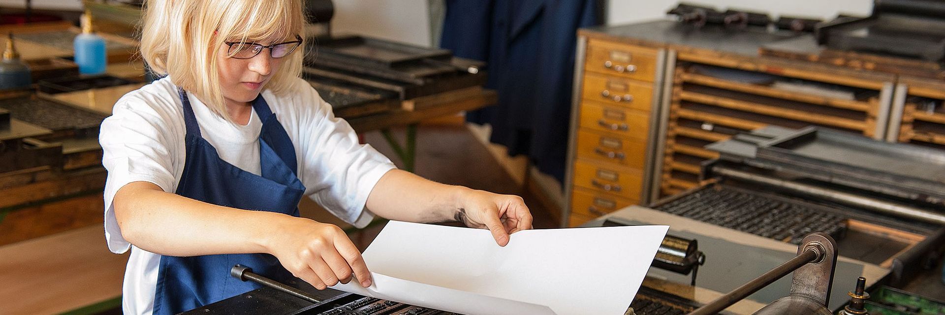 Ein Mädchen steht vor einer kleinen Druckerpresse und legt vorsichtig ein Blatt Papier auf einen gesetzten Text. Sie trägt eine Schürze und hält das Blatt behutsam mit einer Hand an jeder Seite fest.