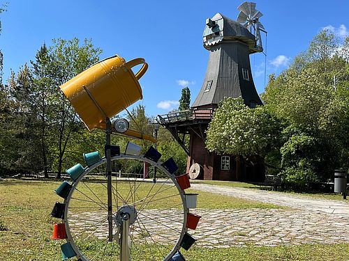Ein Rad mit vielen bunten Bechern steht vor der Bockwindmühle im Museumspark. Über den Bechern ist eine Gieskanne befestigt.
