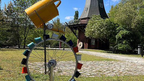 Ein Rad mit vielen bunten Bechern steht vor der Bockwindmühle im Museumspark. Über den Bechern ist eine Gieskanne befestigt.