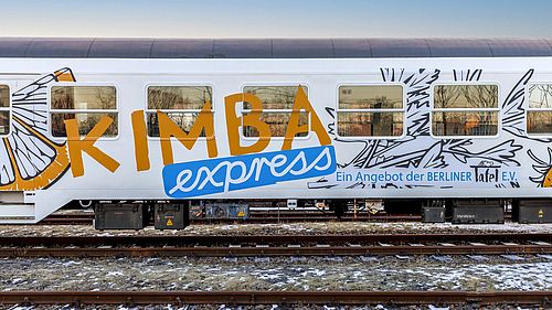 Ein weißer Eisenbahnwagen mit der Aufschrift "KIMBA express" und "Ein Angebot der Berliner Tafel e.V." sowie einer stilisierten Zitronenscheibe und Kräutern.