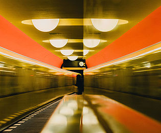 Das Farbfoto zeigt eine U-Bahn-Station mit halbkugelförmigen Deckenleuchten. Auf der linken Seite fährt gerade ein Zug durch. Auf dem zentralen Bahnsteig erkennt man die Silhouette eines Menschen, der unter der Anzeigetafel mit Uhr steht. Die rechte Bildseite wir von einer Fläche eingenommen, auf welcher sich die die Szenerie verschwommen widerspiegelt.