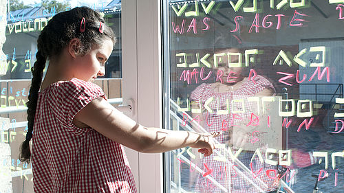 Ein Mädchen deutet auf eine Fensterscheibe, auf der in grün verschiedene Zeichen geschrieben sind. Darunter stehen in roter Farbe Buchstaben und Worte.