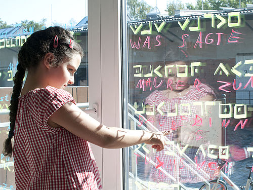 Ein Mädchen deutet auf eine Fensterscheibe, auf der in grün verschiedene Zeichen geschrieben sind. Darunter stehen in roter Farbe Buchstaben und Worte.