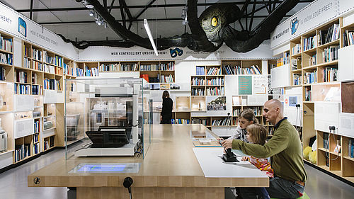 Der Ausstellungsbereich INFORMATION ist wie eine Bibliothek gestaltet. In den vier Wänden sind Regale mit Büchern eingelassen. Im Zentrum steht ein großer Tisch. Darüber schwebt ein riesiger Kraken.