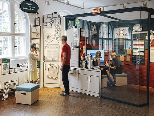 Blick in das gläserne Studio im Ausstellungsbereich "Radio Gleisdreieck". Im Studio sind zwei Personen zu erkennen. Vor dem Studio stehen zwei Menschen und hören etwas über Kopfhörer. Über dem Eingang zum Studio leuchtet der Schriftzug "On Air".