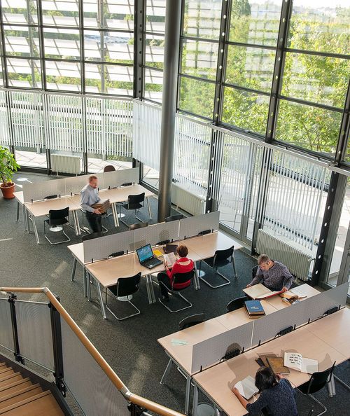 Ein hoher Raum mit Tischen und Stühlen und einem Fensterpanorama mit Blick ins Grüne. An den Tischen sitzen Menschen mit Büchern, Laptops und Schreibutensilien.