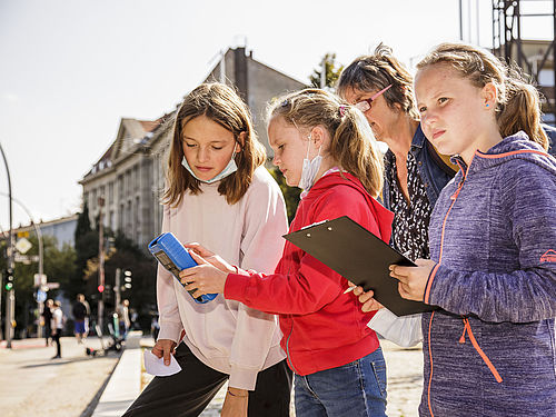 Drei Mädchen stehen auf dem Vorplatz des Museums und halten Tablets in der Hand, auf die sie konzentriert schauen.