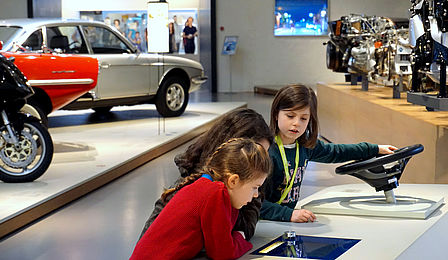 Zwei Mädchen beugen sich über eine Medienstation in der Ausstellung. Daneben schaut ein weiteres Mädchen interessiert hinüber, es hat eine Hand auf ein Lenkrad gelegt. Im Hintergrund sieht man Fahrzeuge und Motoren.