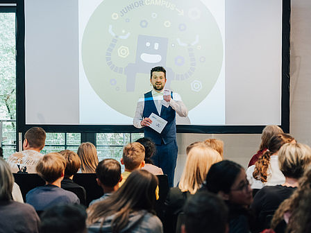 Ein offenbar gut gelaunter Mann im Anzug steht vor einer Leinwand auf der ein lustiger Roboter und die Aufschrift "Junior Campus" zu sehen sind. Vor ihm sitzen Schülerinnen und Schüler in Stuhlreihen und hören ihm gebannt zu.