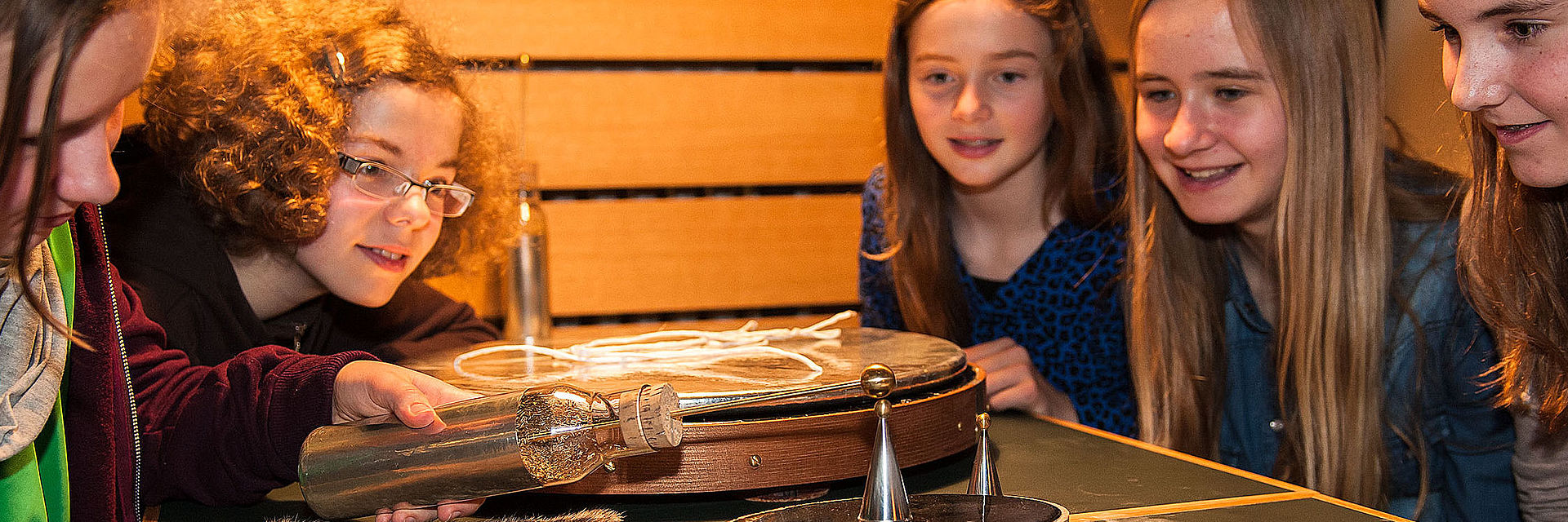 Fünf Mädchen experimentieren an einem Tisch. Ein Mädchen hält eine Glasflasche, aus der ein Stab mit Kugel an der Spitze ragt, an einen kleinen Metallkegel, der auf einer kleinen runden Platte mit glatter, schwarzer Oberfläche steht.