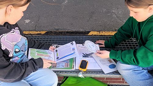 Zwei Kinder sind sitzen auf einer Stufe im Freien mit den Materialen zur GPS-Tour gebeugt. Ein Kind hält eine Verschlüsselungsscheibe in der Hand.