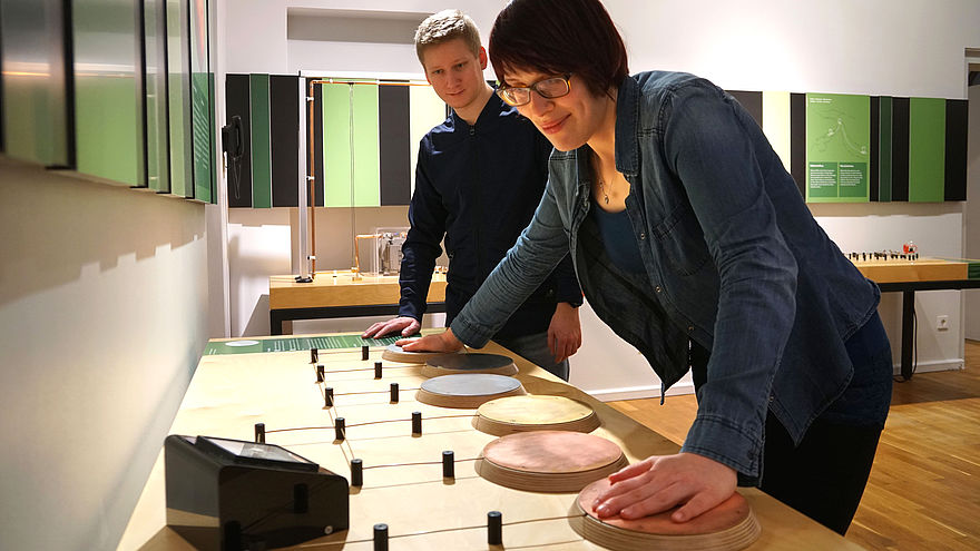 Auf einem Tisch sind verschiedene Metallplatten über Drähte mit einem Messgerät verbunden. Eine Frau berührt zwei der Platten mit ihren Händen und schaut auf das Messgerät, ein Mann schaut dabei zu.