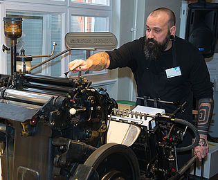 Ein Mann steht an einer großen historischen Druckmaschine. Mit einer Hand betätigt er die Presse, mit der anderen Hand stützt er sich am Rahmen der Maschine ab.
