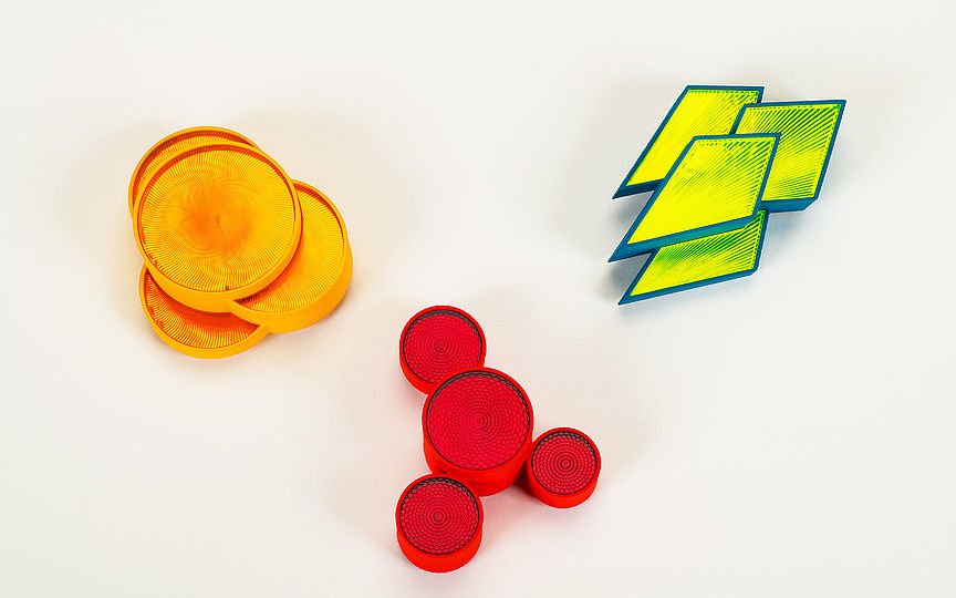Eine orangefarbene, eine rote und eine gelbe Brosche aus Acrylglas. Die Oberflächen sind mit eingravierten Linien verziert.